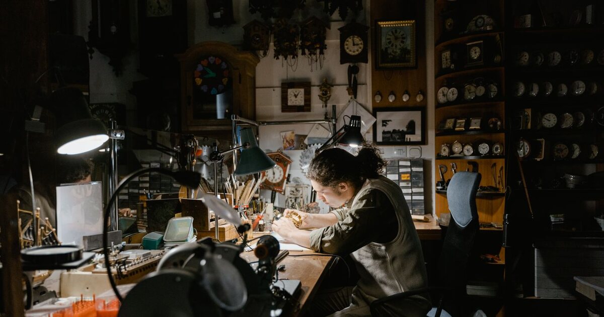 Horloger au travail à son établi dans un atelier de réparation de montres, sous la lumière d'une lampe, entouré de pendules, de réveils et d'outils d'horlogerie