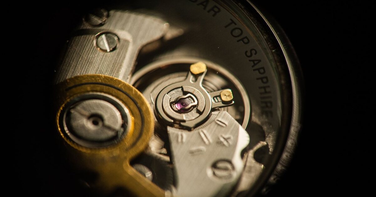 Vue macro du balancier et du spiral d'un mouvement de montre mécanique, avec ses rubis et ses ponts d'acier visibles à travers le fond du boîtier
