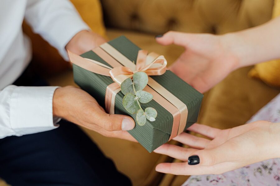 Deux paires de mains échangeant un cadeau emballé de papier vert, ruban rose et brin d'eucalyptus