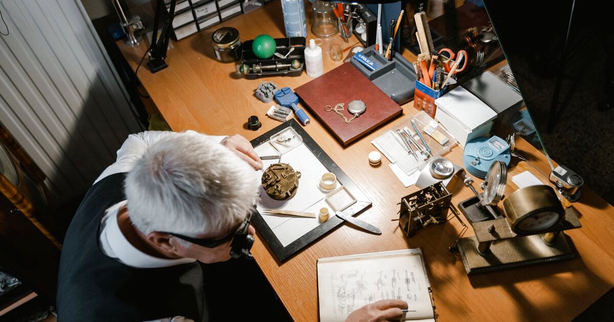 Vue plongeante sur l'établi d'un horloger au travail, avec un mouvement de pendule, des brucelles, une lampe d'atelier et un traité d'horlogerie ouvert