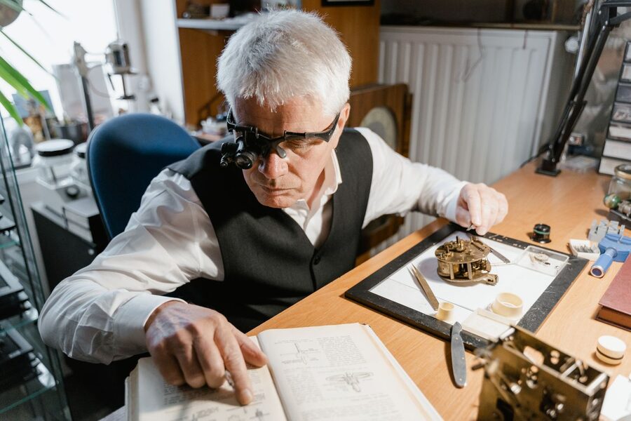 Horloger expérimenté portant des loupes binoculaires, travaillant sur un mouvement à l'établi avec un traité d'horlogerie ouvert devant lui