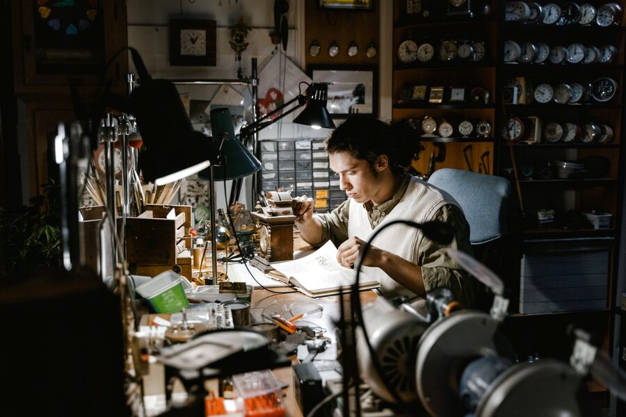Jeune horloger consultant un traité d'horlogerie à son établi, entouré de réveils et de pendules en attente dans un atelier de réparation