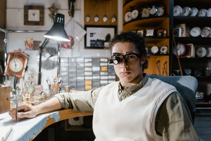 Horloger portant des lunettes loupe binoculaires à son poste de travail, entouré d'établis, de casiers à pièces détachées et de pendules en attente de réparation