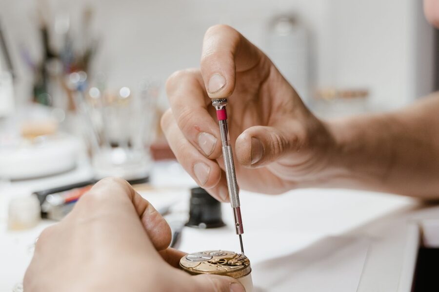 Mains d'un horloger utilisant un tournevis de précision sur un mouvement de montre à l'établi, pièces et outils d'horlogerie à l'arrière-plan