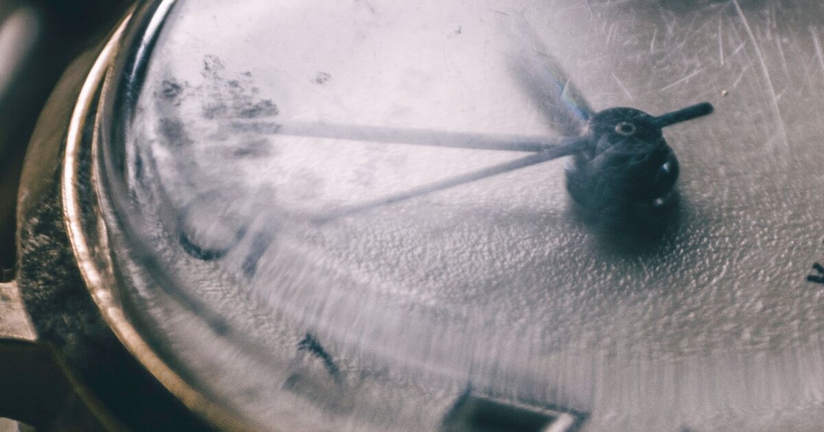 Gros plan sur le verre acrylique bombé d'une montre ancienne, couvert de rayures et devenu laiteux, laissant à peine deviner le cadran et les aiguilles